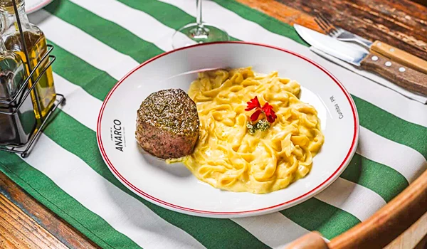 Prato de massa com molho cremoso ao lado de um medalhão de carne, servido em um restaurante com decoração de mesa com toalha listrada verde e branca.