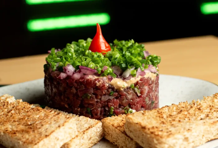 Imagem de um tartar de carne bovina decorado com cebolinhas verdes e uma pimenta vermelha em cima, acompanhado de torradas, representando uma deliciosa receita de tartar de carne.