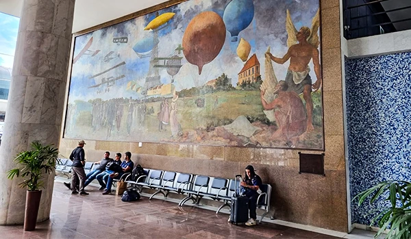 Imagem de um mural artístico na parede de um edifício, com pessoas sentadas em um corredor de aeroporto ou terminal, algumas usando máscaras. Detalhes de detalhes de balões e cenas ao ar livre no mural.
