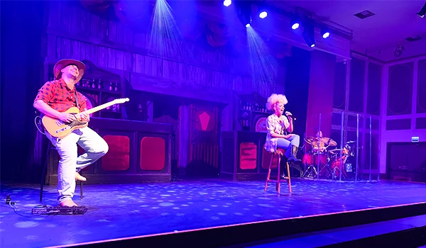 Performers em palco de show musical com iluminação azul, incluindo um guitarrista com chapéu e uma cantora com cabelo branco, ambiente de teatro ou auditório