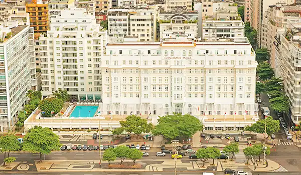 Imagem da fachada do Copacabana Palace, famoso um dos hotéis de luxo no Rio de Janeiro, rodeado por árvores e edifícios altos da cidade.