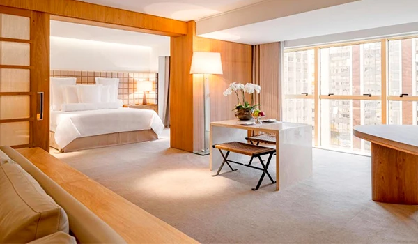 Imagem de um quarto de hotel elegante e bem decorado, com cama confortável, mesa com cadeira de madeira, luminária moderna, e janelas grandes com vista para edifícios na cidade.
