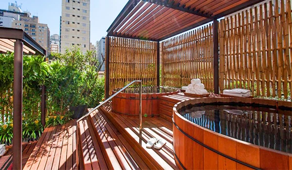 Imagem de uma área de spa ao ar livre com banheiras de hidromassagem de madeira em um prédio na cidade, rodeada por paredes de madeira e plantas verdes, ideal para relaxar na varanda urbana. Um dos hotéis de luxo no Rio de Janeiro.
