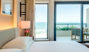 Quarto de hotel com vista para o mar, com cama confortável, luminária e varanda com cadeira e plantas, perfeito para relaxar e apreciar a paisagem marítima