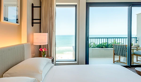 Quarto de hotel com vista para o mar, com cama confortável, luminária e varanda com cadeira e plantas, perfeito para relaxar e apreciar a paisagem marítima