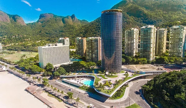 Imagem aérea de um complexo urbano com edifícios modernos e uma montanha verde ao fundo, em um dia ensolarado, destacando o entorno natural e o skyline da cidade.