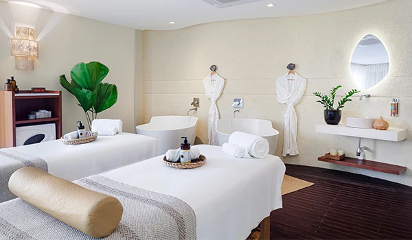 Imagem de uma sala de spa com duas camas de massagem, decoração minimalista, plantas verdes e ambiente tranquilo ideal para relaxamento e bem-estar.
