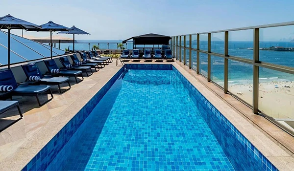 Piscina na cobertura de um hotel com vista para o mar, cadeiras de praia e espreguiçadeiras sob guarda-sóis, ideal para momentos de lazer e relaxamento à beira da água.