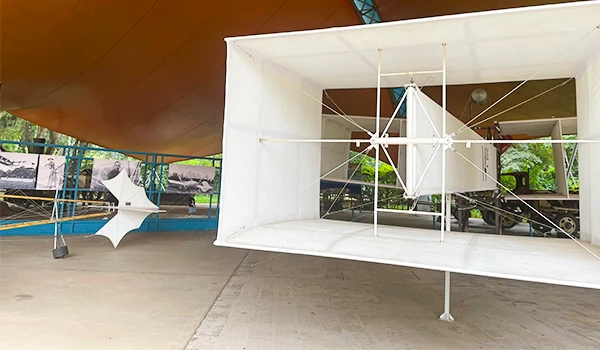 Estrutura de estrutura do avião 14-bis na área ao ar livre com cobertura de tenda laranja e árvores verdes ao fundo. Atrações do Museu Catavento.
