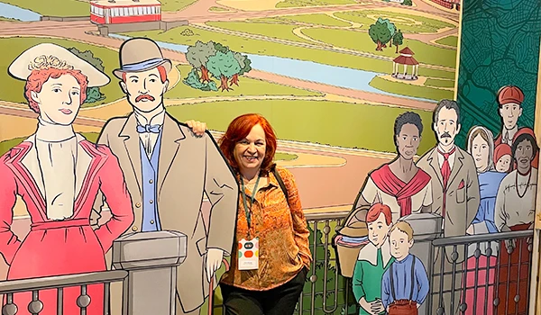 Deusa Rodrigues posando ao lado de figuras de papel de personagens históricos em uma exposição cultural ao ar livre.