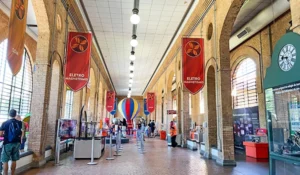 Imagem de uma estação de metrô com banners, balões e visitantes, promovendo o tema do eletromagnetismo, em um ambiente com arquitetura de tijolos expostos.