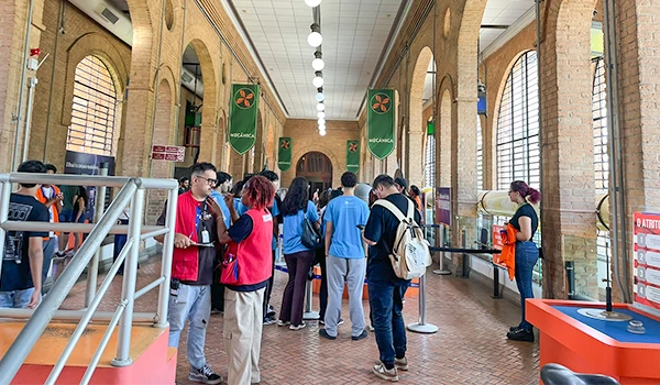 Pessoas formando fila em um ambiente de serviço público ou estação, com arquitetura de tijolos e arcos, algumas com máscaras e mochilas, esperando atendimento ou ingresso. Atrações do Museu Catavento.