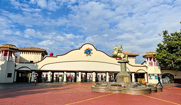 Entrada de parque de diversões com estrutura artística e fonte d'água em primeiro plano, sob céu azul com nuvens, rodeada de árvores.