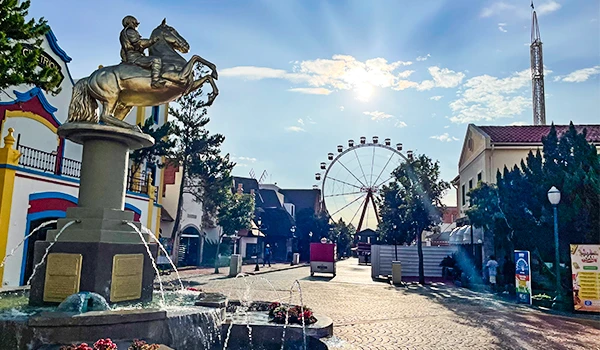 Imagem de uma praça de parque de diversões ao ar livre com roda-gigante, brinquedos e uma estátua de cavaleiro em uma fonte, sob céu azul com algumas nuvens. Ideal para diversão e lazer ao ar livre.