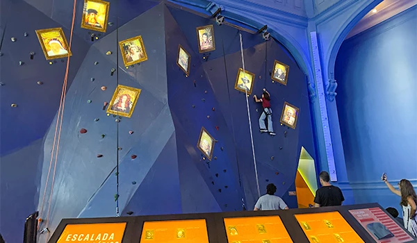 Pessoa praticando escalada em uma parede de escalada indoor, com tela interativa na base para controle e monitoramento.