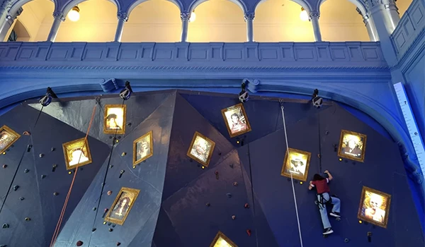 Imagem de uma parede de escalada indoor decorada com quadros dourados suspensos e uma pessoa praticando esportes radicais, em um ambiente com iluminação confortável e detalhes arquitetônicos.