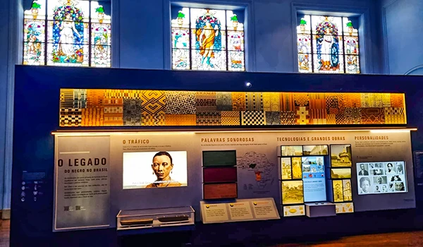 Exposição sobre história no Brasil com vitrais coloridos ao fundo, painel informativo e tecnologia educativa, ideal para projetos escolares.