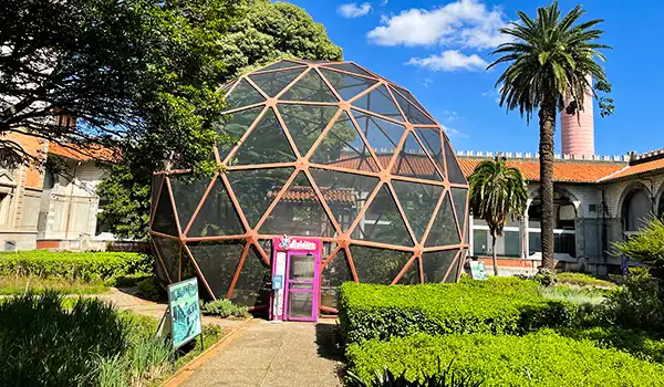 Domo geodésico em um parque com árvores, palmeiras e estrutura histórica ao fundo, sob céu azul com nuvens brancas, promovendo um espaço ecológico e inovador.