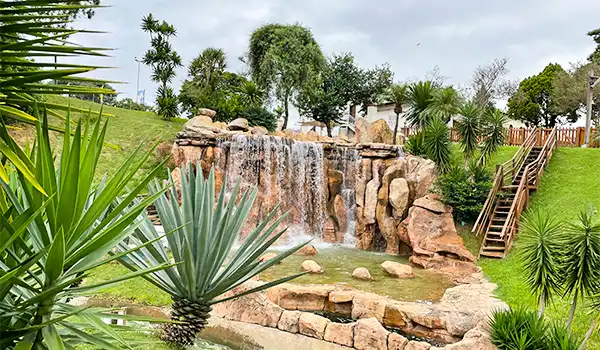 Imagem de uma paisagem de jardim com cascata, vegetação tropical e escadas de acesso, ideal para relaxar ao ar livre.