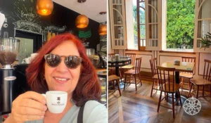 Deusa Rodrigues segurando uma xícara de café em um ambiente de cafeteria, ao lado de uma sala com mesas e cadeiras próximas a uma janela com vista para o jardim.