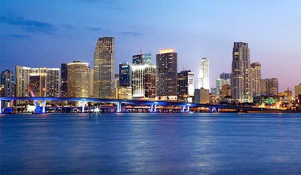 Foto de Miami ao pôr do sol, com seus prédios ao fundo e a ponte e o mar em primeiro plano. Cidades dos Estados Unidos na Copa.