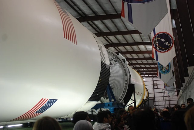 Interior de um hangar com uma aeronave grande ao lado de várias bandeiras penduradas, vista de perto da fuselagem e do motor com janelas e estrutura metálica no teto.
