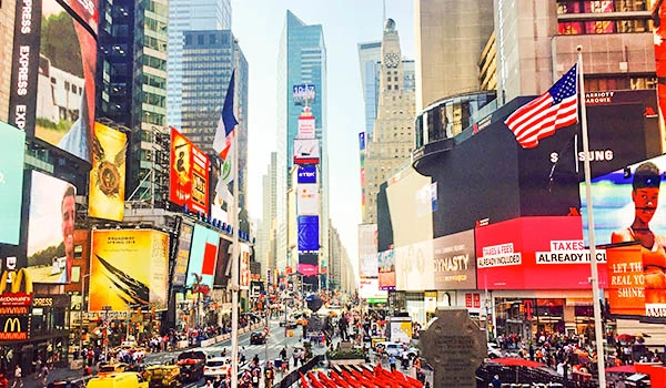 Vista de Times Square em Nova York com arranha-céus, anúncios luminosos nas fachadas, faixa dos Estados Unidos e multidão circulando na rua movimentada.