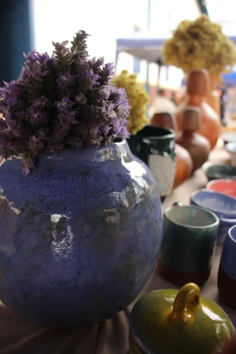 Vaso de cerâmica azul com flores roxas em primeiro plano, com fundo desfocado mostrando mesa decorada e objetos de decoração em ambiente interno. Cunha Capital da Cerâmcia.
