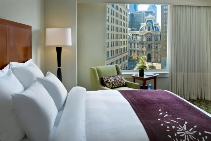 Quarto de hotel moderno com cama de casal bem arrumada, edredom roxo, almofadas brancas, poltrona verde e janela com vista para prédios altos e skyline urbano ao fundo.
