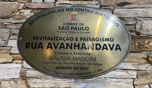 Placa oval de metal fixada em uma parede de pedras, com informações sobre a revitalização do centro histórico de São Paulo. O texto destaca “Revitalização e Paisagismo Rua Avanhandava”, além de mencionar a Prefeitura de São Paulo, o prefeito Gilberto Kassab, cantinhos charmosos de SP.