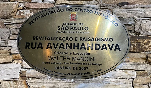 Placa oval de metal fixada em uma parede de pedras, com informações sobre a revitalização do centro histórico de São Paulo. O texto destaca “Revitalização e Paisagismo Rua Avanhandava”, além de mencionar a Prefeitura de São Paulo, o prefeito Gilberto Kassab, cantinhos charmosos de SP.