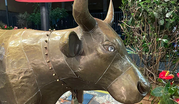 Escultura metálica de um boi ou touro em tom bronze, vista de perfil e em primeiro plano. O animal tem dois chifres, olhos claros e detalhes de rebites e emendas na superfície. Ao fundo, há vegetação, flores e parte de uma construção.