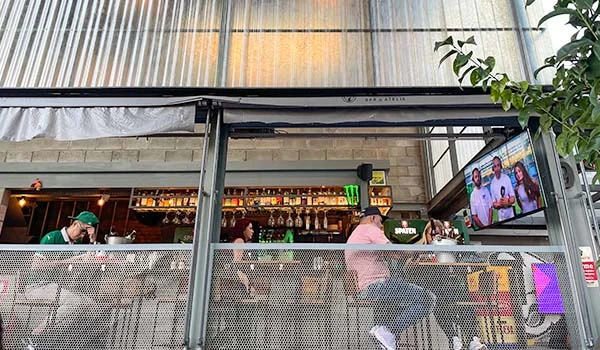 Foto do Bar Ateliê e Brotero 39 com fachada com vidro, onde se observa o balcão com bebida, clientes nas mesas e uma televisão ligada. Uma das coisas legais para fazer na Barra Funda.