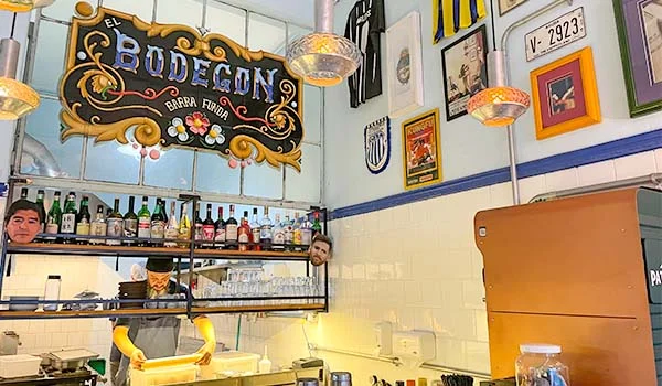 Foto interna do El Bodegón Barra Funda bar e restaurante, estilo argentino. Onde se observa a placa do estabelecimento, funcionário trabalhando, garrafas, quadros na parede e uma máscara do jogador Maradona, na Barra Funda.