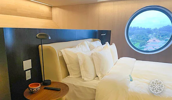 Quarto de hotel com cama de casal, lençóis e travesseiros brancos, cabeceira escura, luminária de leitura, mesa lateral de madeira e janela circular com vista para a paisagem externa.