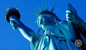 Estátua da Liberdade, em Nova York, vista de baixo para cima, com a tocha erguida e a coroa com raios destacados contra o céu azul.