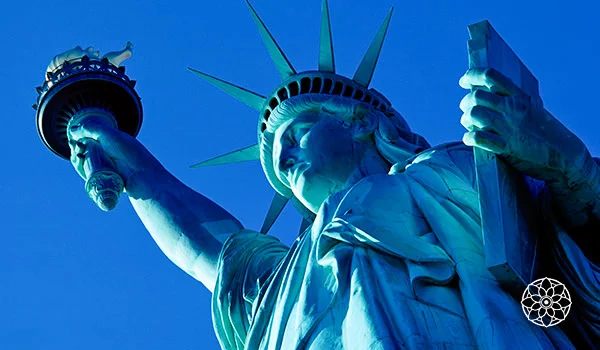 Estátua da Liberdade, em Nova York, vista de baixo para cima, com a tocha erguida e a coroa com raios destacados contra o céu azul.