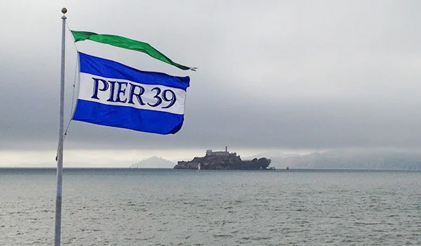 Foto de uma bandeira azul, branca e verde com os dizeres, Pier 39, ao fundo a Ilhas de Alcatraz e o tempo nublado em São Franciso, Estados Unidos.