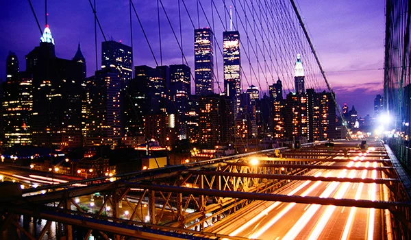 Foto noturna da Ponte do Brooklyn em Nova York com vários prédios iluminados em volta.