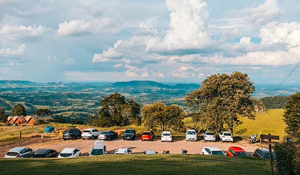 Vista panorâmica de uma paisagem rural montanhosa, com céu azul e muitas nuvens, árvores e campos verdes ao fundo. Em primeiro plano, há um estacionamento com vários carros e algumas construções.