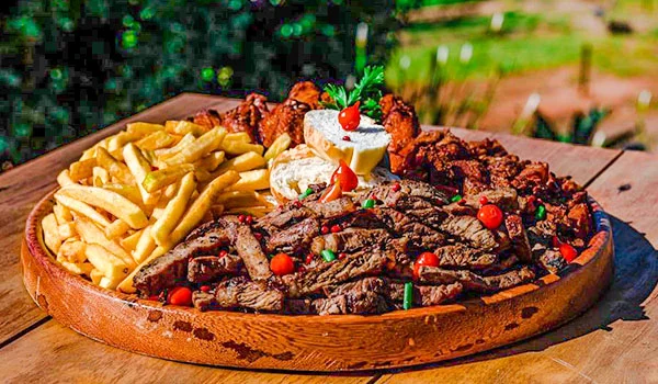 Prato servido em uma tábua de madeira, com tiras de carne grelhada, batatas fritas, pedaços de queijo e tomates-cereja, decorado com ervas frescas. A refeição está ao ar livre, sobre uma mesa de madeira, com vegetação ao fundo.