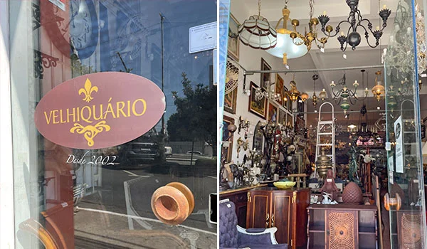 Vitrine do antiquário Velhoquário, identificado pela placa “Desde 1992”, com reflexo da rua no vidro. Ao lado, o interior da loja exibe móveis antigos, objetos decorativos, luminárias, lustres e peças de antiquário.