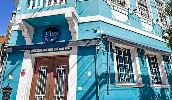 Fachada azul-turquesa de um bar no Bixiga, com portas e janelas protegidas por grades brancas e uma placa circular com o nome “Blue Bar”. O que fazer no Bixiga.