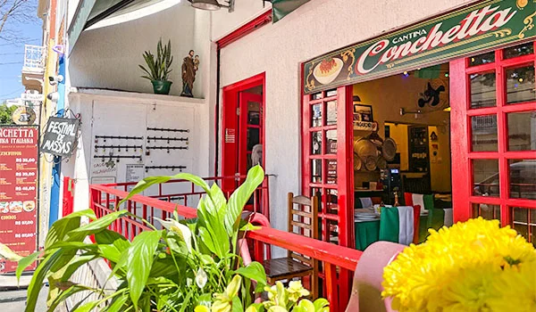 Fachada de uma cantina italiana com portas e janelas vermelhas, placa com o nome “Cantina Conchetta”, mesas e cadeiras na entrada e plantas em vasos.