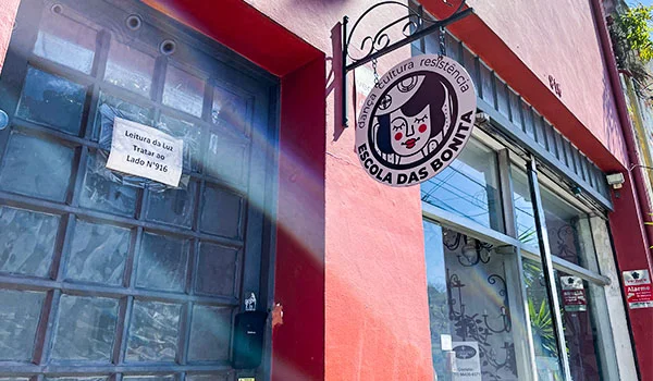Fachada vermelha e branca com uma placa pendurada indicando “Escola das Bonita”, acompanhada de um desenho de um rosto feminino. Na porta, há um aviso sobre “Leitura das Luz”