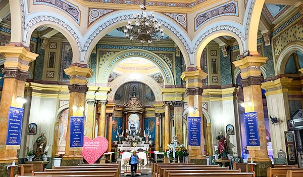 Interior ornamentado de uma igreja, com altar ao fundo, imagens religiosas, colunas decoradas, arcos pintados e bancos de madeira para os fiéis. O que fazer no Bixiga.
