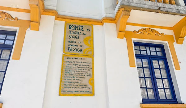 Detalhe de uma fachada histórica branca e amarela com placa informativa que diz “Respeite a história do Bixiga” e apresenta informações sobre a preservação do bairro. O que fazer no Bixiga.