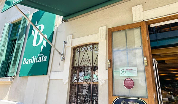 Foto em montagem com duas imagens. À esquerda, uma mulher de cabelos ruivos, usando blusa verde-clara e colar, sorri enquanto leva um pedaço de comida à boca com um garfo, em um restaurante. À direita, uma placa emoldurada na parede informa que o restaurante Basilicata foi finalista na categoria Trattoria do prêmio Veja São Paulo Comer & Beber 2023/2024.