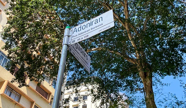 Placas de rua indicando “Adoniran Barbosa” e “Rua Adoniran Barbosa”, instaladas em um poste sob a copa de árvores. O que fazer no Bixiga.