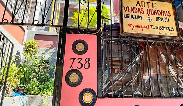 Entrada de um imóvel com fachada rosa, grades pretas e três discos decorativos na parede. Uma placa anuncia “Arte, vendas, quadros, Uruguay, Brasil” e identifica o endereço pelo número 738.
